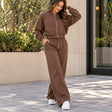 Woman in a brown cropped zip jacket and matching wide leg jogging pants outside a stone building with green planters