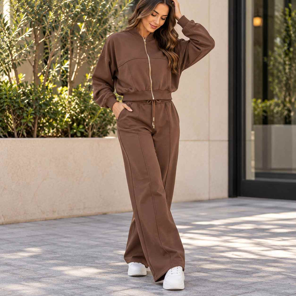 Woman in a brown cropped zip jacket and matching wide leg jogging pants outside a stone building with green planters