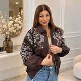 Woman zipping a chocolate brown bomber jacket with white beaded flowers over a crop top and jeans