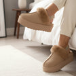 Brown fuzzy slippers with fur trim worn by a person sitting on a bed.