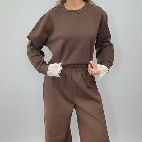 Brown sweatshirt and joggers with hands in the pockets, lace peeking at the cuffs