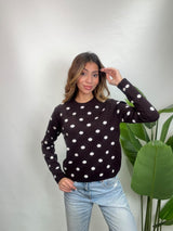 Woman in a brown white polka dot sweater touching her hair, standing beside a tall plant