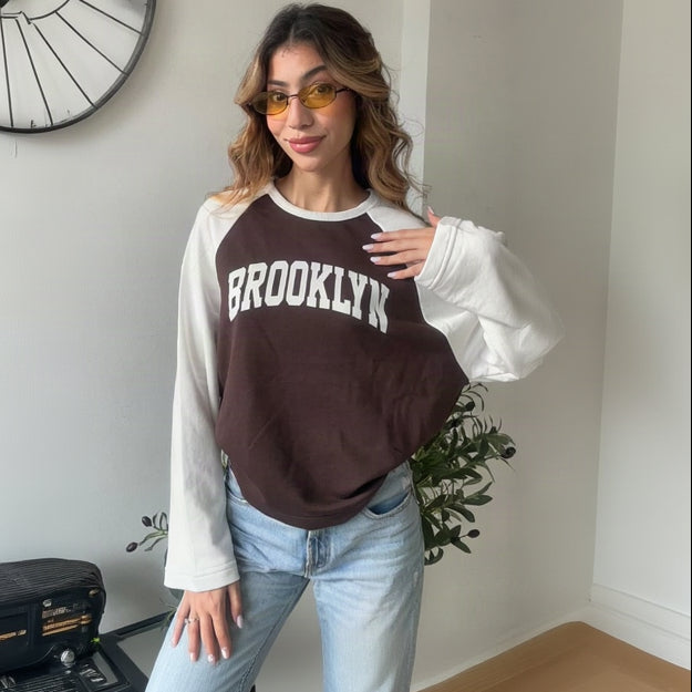 Woman wearing a brown and white raglan Brooklyn sweatshirt with light wash jeans