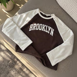 Brown Brooklyn sweatshirt with white raglan sleeves laid flat on a grey sofa
