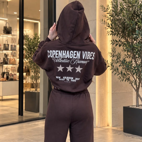 Back of a brown cropped hoodie with a large slogan graphic and white stars, worn with wide leg pants