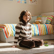 Boy sitting on a rug in a brown and white striped half zip pullover and brown joggers
