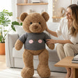 Curly brown teddy bear wearing a charcoal grey ribbed jumper printed with a blush cheeked bear face