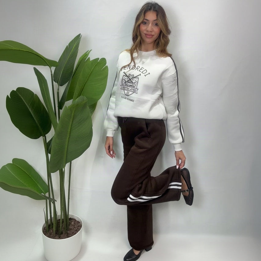 Woman posing with one knee raised in a white varsity sweatshirt and brown side stripe pants