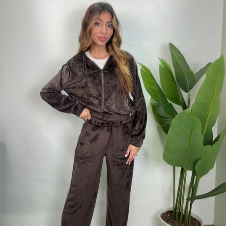 Woman in a brown velour zip hoodie and wide leg pants set standing beside a green plant