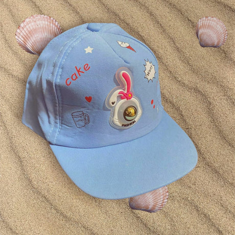Baby blue kids cap angled on sand, bunny patch and small heart, star and cupcake doodles