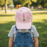 Pale pink kids baseball cap with a raised bunny patch and doodle prints, worn by a girl in dungarees