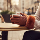 A person seated at an outdoor cafe table wearing burgundy leather gloves with a fluffy orange-brown faux fur trim, holding a steaming white cup