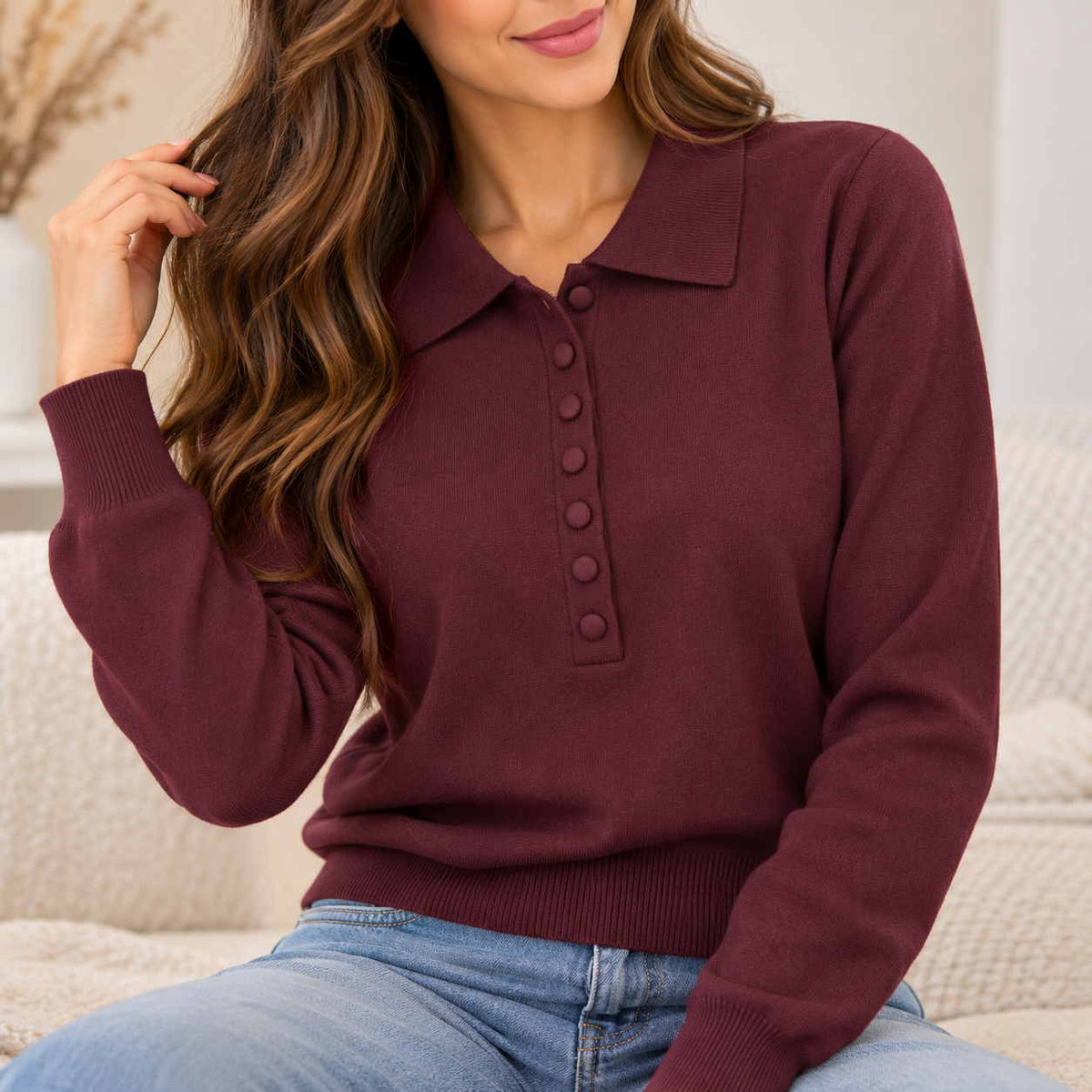 Close up of a woman in a burgundy knit polo sweater with covered buttons, worn with light blue jeans on a cream sofa