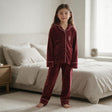 Girl standing barefoot in a burgundy velvet pyjama set with pink piping beside a bed
