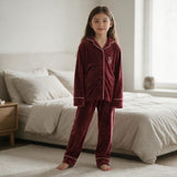Girl standing barefoot in a burgundy velvet pyjama set with pink piping beside a bed