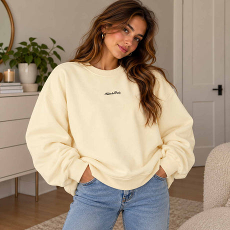 Woman in a butter yellow crew neck sweatshirt with a dainty Aube de Paris script, hands in her jeans pockets