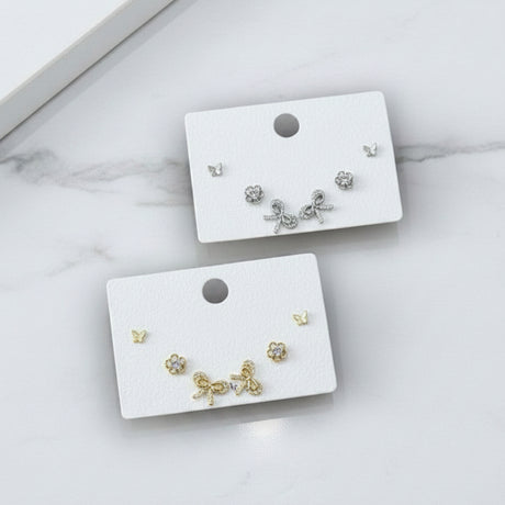 Silver and gold set of butterfly bow stud earrings on white display cards against a marble background