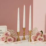Three short gold holders with white tapers beside roses and scattered petals
