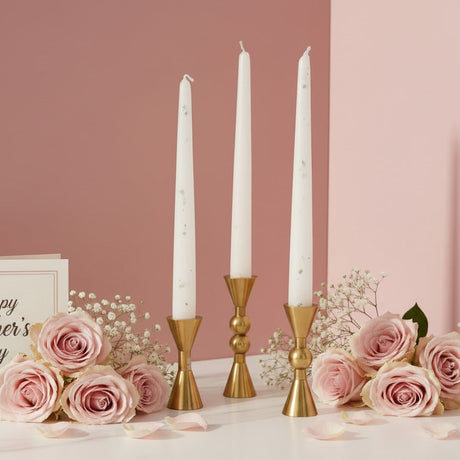 Three short gold holders with white tapers beside roses and scattered petals