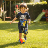 Boy running after a ball outdoors in a navy set with a surfing dinosaur graphic tee and shorts