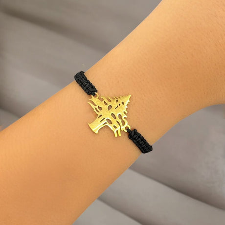 Gold cedar pendant on a black cord bracelet worn on a wrist against a neutral background