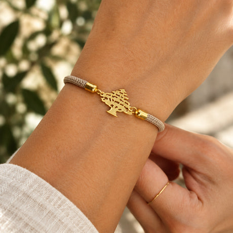 Wrist wearing a beige braided cord bracelet with a gold cut out cedar tree charm