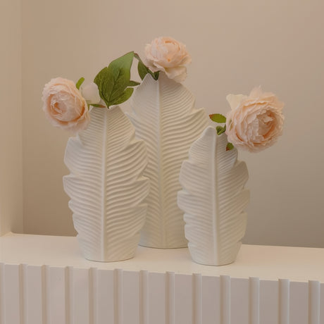 Three matte white leaf shaped vases on a fluted console, each holding a single blush peony