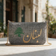 Heathered charcoal grey pouch with Lebanon in gold Arabic and a green cedar, in front of an old house with green shutters