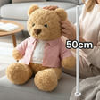 Curly caramel teddy bear in an open pink shirt seated on a grey sofa, marked 50cm