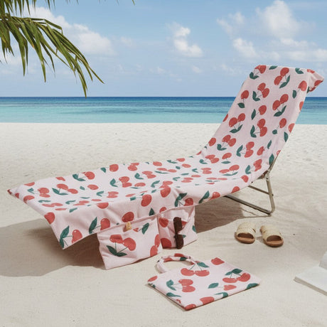 A beach lounge chair and matching towel set with a pink cherry print, placed on white sand at the beach with ocean, sky, and palm leaves visible.