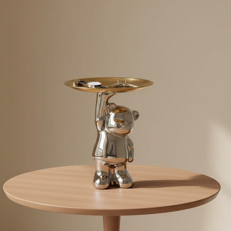 Metallic bear-shaped object with a tray on a wooden table against a beige background