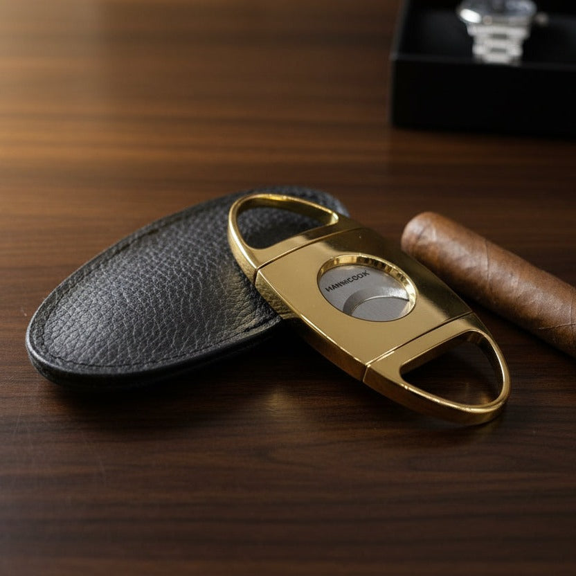Gold cigar cutter with black leather case and brown cigar on wooden surface
