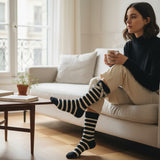 Woman sitting on a couch wearing black and white srtiped bow socks holding a mug in a cozy living room