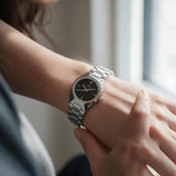 Silver steel watch with black dial worn on the wrist by a window