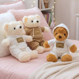 Three tracksuit teddy bears with gold Love chest patches arranged on pink bedding