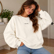 Woman in a cream crew neck sweatshirt with a dainty Aube de Paris script, hands in her jeans pockets