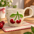 Glossy red cherries on a green stem sculpted onto a cream mug, fresh cherries beside it