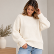 Woman in a cream chunky wool knit sweater and light jeans touching her hair beside a vase of dried stems