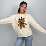 Cream knit sweater with a brown teddy bear in a red party hat and bow, arms out
