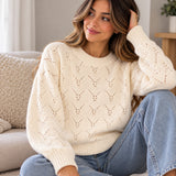 Cream pointelle crew neck sweater with ribbed cuffs, worn sitting on a sofa with jeans