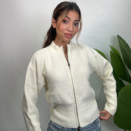 Cream ribbed knit zip up cardigan zipped to the funnel neck, worn with light jeans by a plant