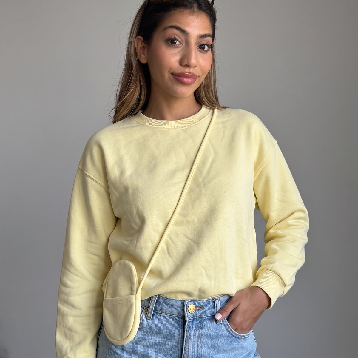 Pastel yellow sweatshirt with a tonal crossbody pouch at the hip, grey studio backdrop