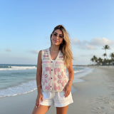 Woman in a pink patterned vest and white shorts standing on a beach with palm trees in the background.