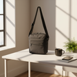 Grey crossbody bag on a white table with a cup and notebook in a bright room