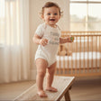 Baby standing on a wooden bench in an ivory short sleeve bodysuit printed with a Daddys Main Squeeze slogan