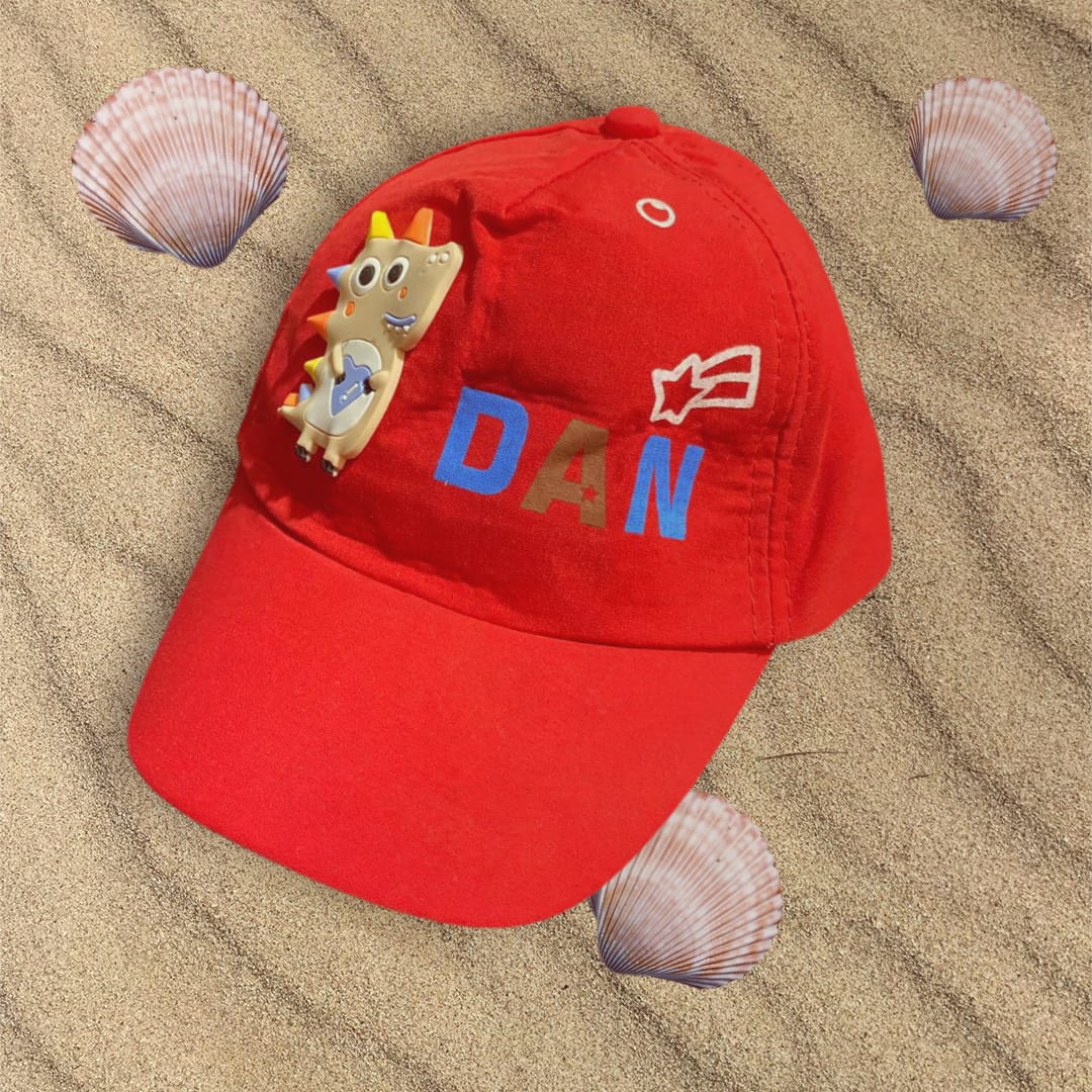 Red cotton boys cap lying flat on sand with a soft dinosaur patch and star motif