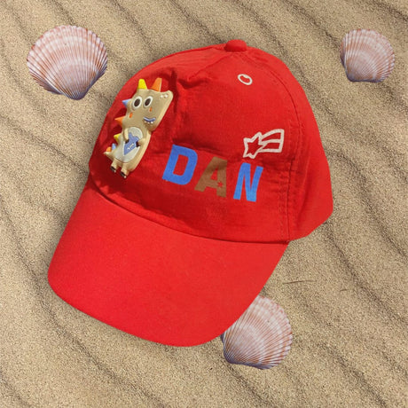Red cotton boys cap lying flat on sand with a soft dinosaur patch and star motif