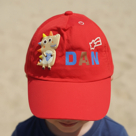 Red kids baseball cap with a rubber dinosaur patch and blue lettering, worn by a young boy