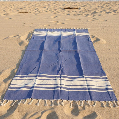 Dark blue and white striped beach towel with tassel edges on sandy beach with ocean view
