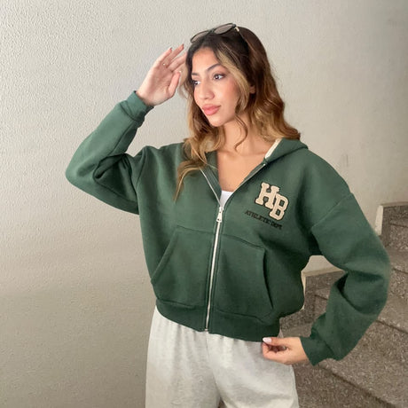 Woman in a dark green cropped zip hoodie with a beige letter patch and sunglasses on her head